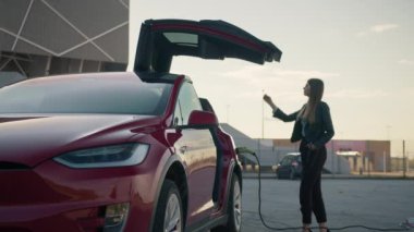 Young beautiful Stylish girl approaches a luxurious modern electric car, which is charged at the charging station. The girl closes the car door with a button, the modern door closes automatically.