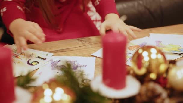 暖炉とクリスマスツリーの近くのテーブルに少女が描かれています サンタの手紙を持っている子供 幸せな女の子は部屋の中で新年の木によってサンタのための写真を描く — ストック動画