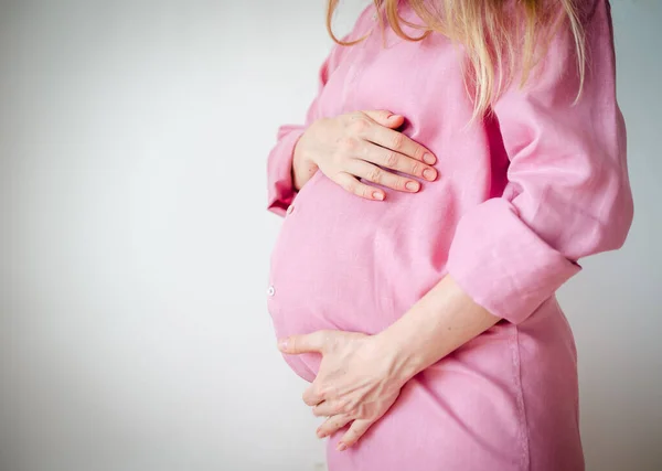 Mulher grávida em uma camisa rosa segura as mãos em seu estômago em um fundo branco — Fotografia de Stock