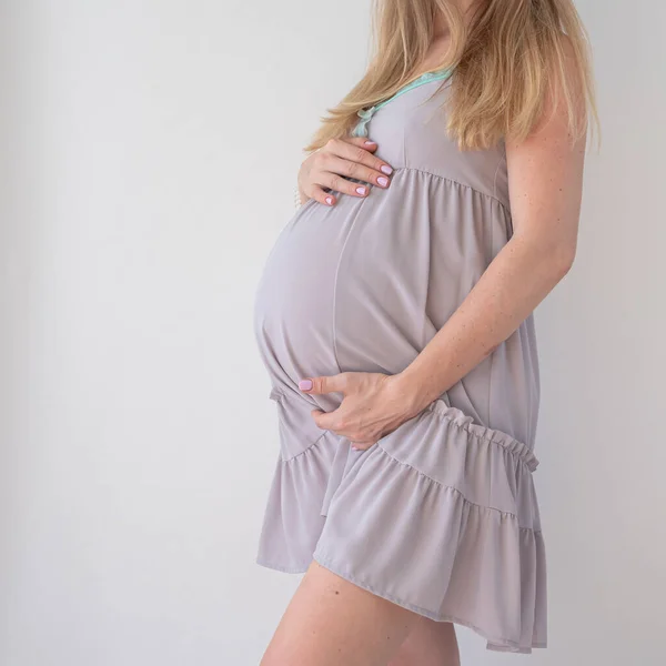 Uma mulher grávida em uma camisola lilás pálida fica em um fundo branco e segura as mãos em seu estômago — Fotografia de Stock