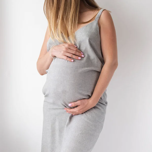 Een verfijnde zwangere vrouw in een grijze jurk staat op een witte achtergrond met haar handen op haar buik, het concept van vrouwelijkheid — Stockfoto