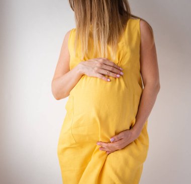  Sarı elbiseli hamile bir kadın beyaz bir arka planda duruyor. Elleri karnında. Kadınlık kavramı.