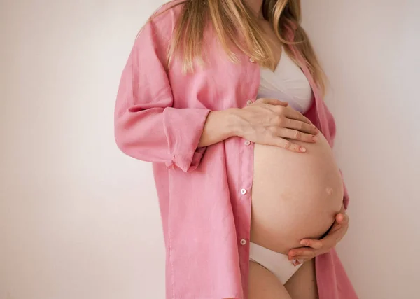 Una mujer embarazada tercer trimestre en ropa interior y una camisa rosa se coloca sobre un fondo blanco — Foto de Stock