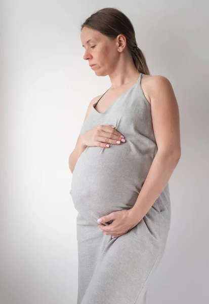 Donna incinta in abito grigio sta su uno sfondo bianco con le mani sullo stomaco, il concetto di femminilità — Foto Stock