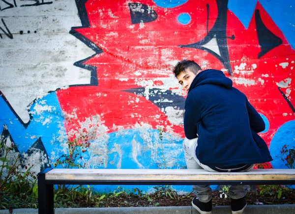 Young man with hoodie sitting on bench in front of graffiti — Stock Photo, Image