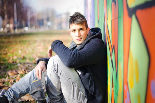 Handsome young man sitting on ground against colorful graffiti — Stock Photo, Image