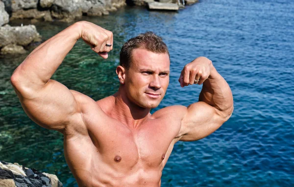 Double biceps pose for young bodybuilder by the sea — Stock Photo, Image