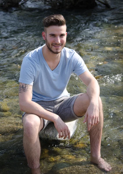 Handsome young man sitting on river rock — Stock Photo, Image
