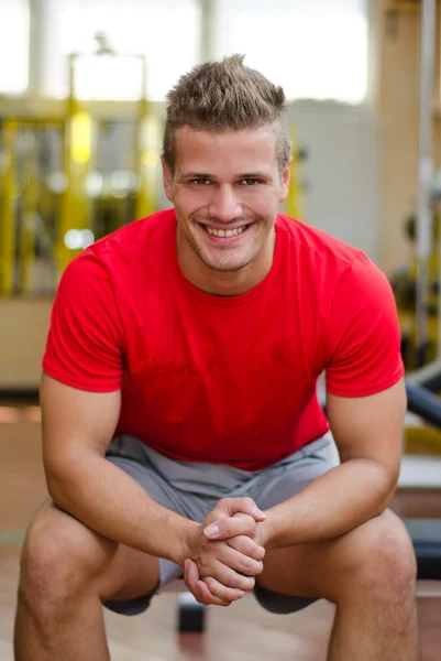 Attractive young man in gym sitting on bench, smiling — Stock Photo, Image