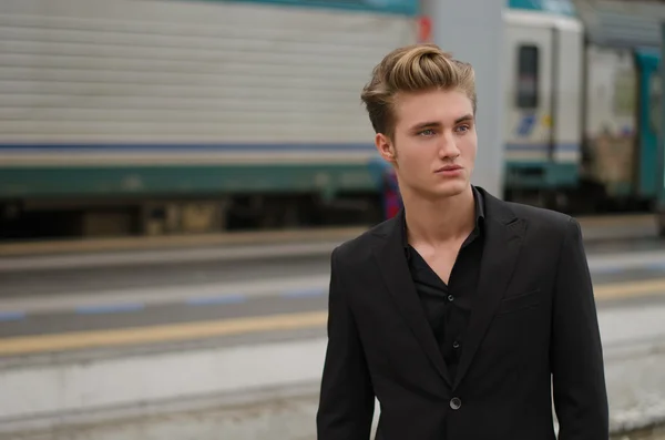 Handsome blond young man with train behind him, in railway station — Stock Photo, Image