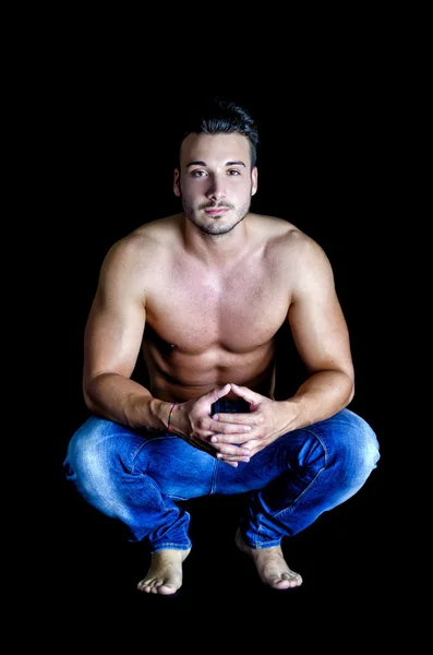 Shirtless muscular young man in jeans sitting on dark background — Stock Photo, Image