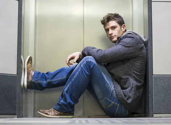 Jovem bonito sentado em frente de portas de elevador — Fotografia de Stock