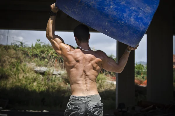 Back of muscular construction worker shirtless in building site holding big barrel — Stock Photo, Image