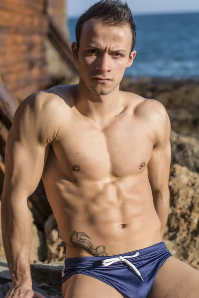 Musculoso joven sin camiseta tomando el sol en la roca por el mar —  Fotos de Stock