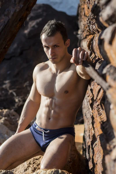 Muscular jovem sem camisa, tomando banho de sol na rocha por parede de metal enferrujada — Fotografia de Stock
