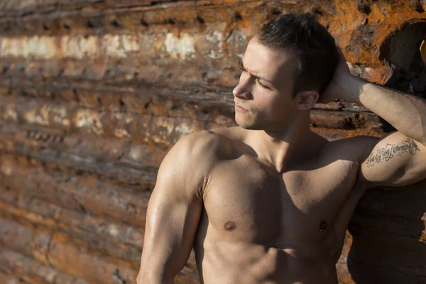 Musculoso joven sin camiseta tomando el sol en la roca por la pared del metal oxidada —  Fotos de Stock