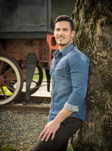 Sonriente a joven de camisa vaquera contra árbol cerca tren viejo —  Fotos de Stock