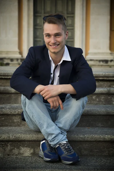 Smiling blond young man sitting on stone stair steps outside — Stock Photo, Image