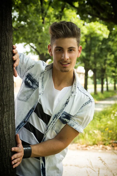 Friendly handsome young man behind tree smiling — Stock Photo, Image