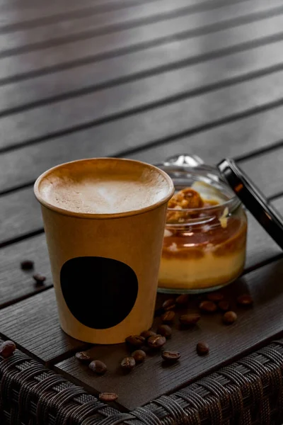 在纸杯中倒入咖啡 在木制桌子上的玻璃瓶中加入盐焦糖的美味甜点 有选择的重点 柔和模糊的背景 顶部视图 免版税图库照片