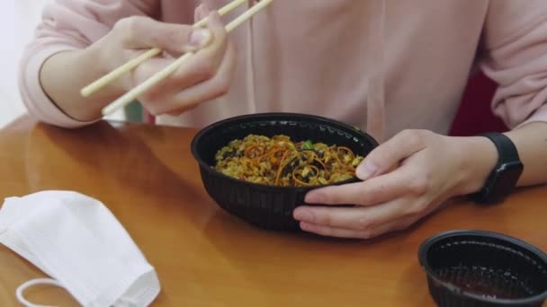 La ragazza mangia tagliatelle asiatiche con pollo, verdure e salsa agrodolce con bastoncini di legno seduti in un fast food, sul tavolo c'è una maschera medica protettiva.. — Video Stock