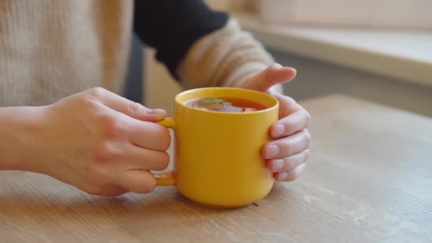Vrouw houdt haar handen op een gele mok hete citrus thee in afwachting van haar vriendje in een cafe. — Stockvideo