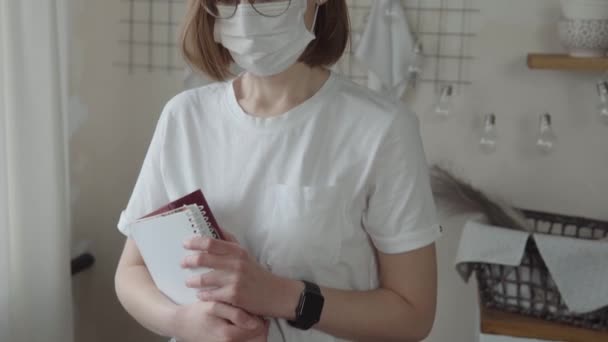Egy szemüveges fiatal nő portréja, védőorvosi maszk, fehér póló könyvekkel a kezében.. — Stock videók