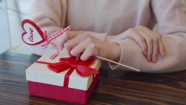 Dziewczyna przed podarowaniem prezentu kochankowi trzyma w dłoni wystrój w postaci serca. Walentynki, rocznica, koncepcja. Na stole jest prezent w czerwono-białym pudełku.. — Wideo stockowe