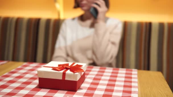 Dziewczyna rozmawia przez telefon przed randką w ciemno w kawiarni. Pudełko prezentów w kolorze czerwono-białym z kokardą na pierwszym planie. Koncepcja złej randki, miłości i związków. — Wideo stockowe