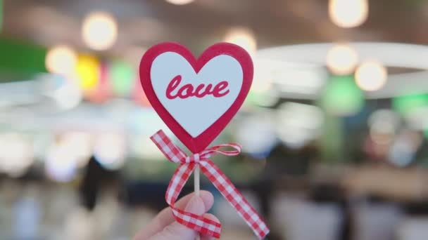 Symbol der Liebe ist ein herzförmiger Stock in der Hand auf dem Hintergrund eines öffentlichen Platzes in einem Einkaufszentrum und eines Restaurants auf dem Food Court. Das Konzept des Valentinstags-Feiertags. — Stockvideo