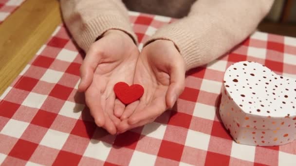 Дівчина в коханні показує червоне серце в своїх долонях як символ любові. Подарунок у пачці у формі серця на столі. Романтичний момент на побаченні, оголошення кохання або пропозиція про шлюб . — стокове відео