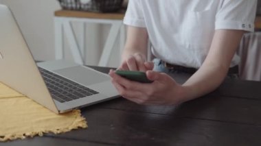 Akıllı telefondan mesaj yazan modaya uygun bir kadın, merkez ofiste modern dizüstü bilgisayarın önünde oturuyor. Yakın çekim. Yüksek kalite 4k görüntü.