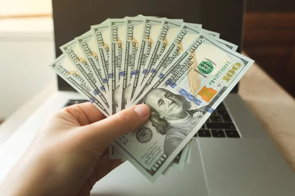 A man holds a fan of hundred dollar bills in his hand against the background of a modern laptop.
