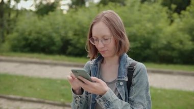 Student woman in glasses denim jacket chatting online, uses smartphone social media, paying online bills, shopping in web. Slow motion footage.