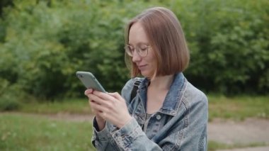Cheerful girl student 20s 30s in glasses chatting online, uses smartphone social media, paying online in web, shopping. Slow motion footage.
