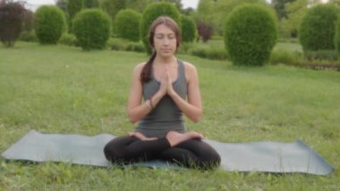 Front view of a woman instructor sitting on yoga mat in lotus position outside in city park. Namaste mudra, seated yoga asana, lotus pose, lotus posture, padmasana, cross legged sitting posture.