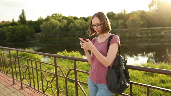 Smiling woman in glasses with a leather backpack using smartphone standing outdoors on the bridge. Hipster girl browsing Internet on a phone, texting and communicating.
