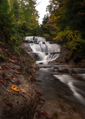 Sonbaharda Yukarı Michigan 'daki Sable Falls' da, kıyı şeridini, nehir taşlarını ve ön plandaki kayaları kirleten yapraklarla birlikte güzel, uzun pozlu bir fotoğraf..