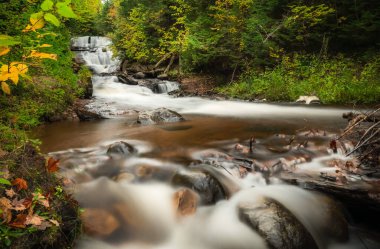 Sonbaharda Yukarı Michigan 'daki Sable Falls' da, kıyı şeridini, nehir taşlarını ve ön plandaki kayaları kirleten yapraklarla birlikte güzel, uzun pozlu bir fotoğraf..