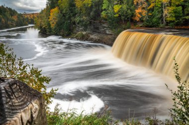 Michigan 'daki Yukarı Tahquamenon Şelalesi' nin sonbahar panoramik fotoğrafı. Suyun çağlayarak nehre döküldüğü ve etrafının yemyeşil ağaçlarla çevrili olduğu ve ön planda yeşillik ve ahşap direkli renkli yapraklarla çevrildiği yer..