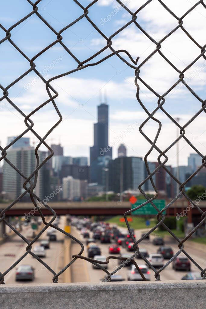 An out of focus shot of the downtown Chicago skyline framed by a chain link fence with an opening in it in focus in the foreground.