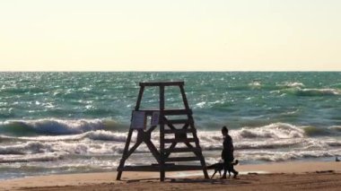 Chicago, IL - July 9th, 2022: A woman walks her dog on the sand along Foster Beach past an empty lifeguard tower in the morning as waves crash into the shoreline from Lake Michigan.