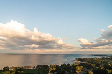 Gün batımında mavi gökyüzünde pofuduk beyaz ve sarı kümülüs bulutlarıyla Michigan Gölü 'nün güzel panoramik manzarası sakin sulardan yansıyor. Sıcak bir yaz gününde insanlar plajın keyfini çıkarırken..