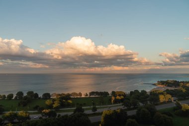 Gün batımında mavi gökyüzünde pofuduk beyaz ve sarı kümülüs bulutlarıyla Michigan Gölü 'nün güzel panoramik manzarası sakin sulardan yansıyor. Sıcak bir yaz gününde insanlar plajın keyfini çıkarırken..