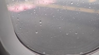 Handheld close up shot of rain collecting on an airplane window during inclement weather as the plane moves across the runway for takeoff.