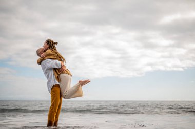 Kumsalda oynaşan genç bir çift, havada bir kızı tutan bir adam. Görüntü, çift ilişkisinin samimiyetini gösteriyor. Yüksek kalite fotoğraf. Bulutlu hava.