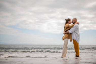 Genç ve sevgi dolu bir çift sahilde ayakta durup sarılıyor. Görüntü, çift ilişkisinin samimiyetini gösteriyor. Görünüşe göre o çift öpüşmek üzere. Yüksek kalite fotoğraf. Bulutlu hava.