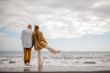 Genç ve sevgi dolu bir çift sahilde ayakta durup sarılıyor. Görüntü, çift ilişkisinin samimiyetini gösteriyor. Görünüşe göre o çift öpüşmek üzere. Yüksek kalite fotoğraf. Bulutlu hava.