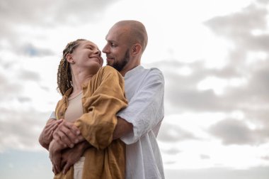 Genç ve sevgi dolu bir çift sahilde ayakta durup sarılıyor. Görüntü, çift ilişkisinin samimiyetini gösteriyor. Görünüşe göre o çift öpüşmek üzere. Yüksek kalite fotoğraf. Bulutlu hava.