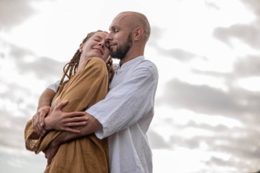 Genç ve sevgi dolu bir çift sahilde ayakta durup sarılıyor. Görüntü, çift ilişkisinin samimiyetini gösteriyor. Görünüşe göre o çift öpüşmek üzere. Yüksek kalite fotoğraf. Bulutlu hava.